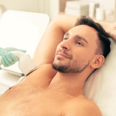 Smiling young man lying on the pillow with one hand behind his head and having laser hair removal on his armpit